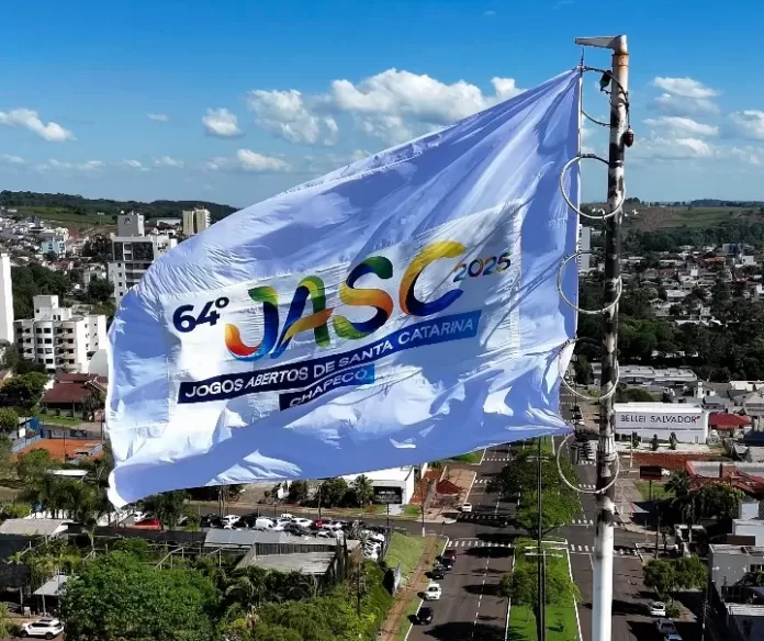 Grande bandeira dos Jogos Abertos de Santa Catarina hasteada no mastro da prefeitura de Chapecó, com céu azul ao fundo