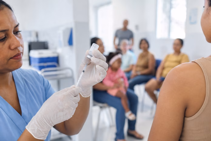 Profissional de saúde prepara vacina enquanto pacientes aguardam atendimento em uma unidade de vacinação