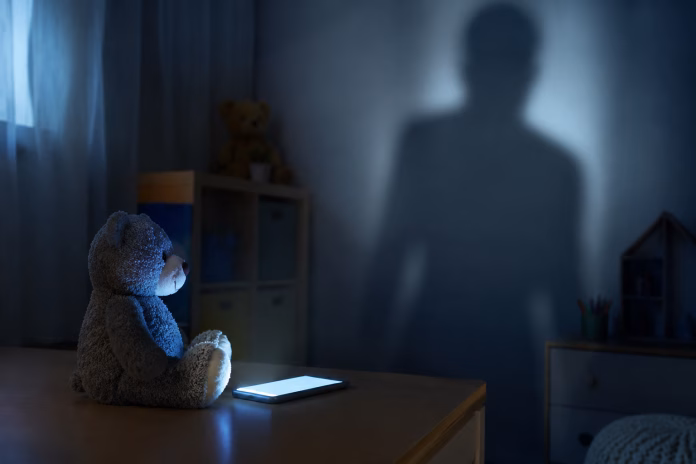 Urso de pelúcia diante de um celular iluminado, enquanto a sombra de um adulto na parede simboliza os riscos do aliciamento infantil na internet.